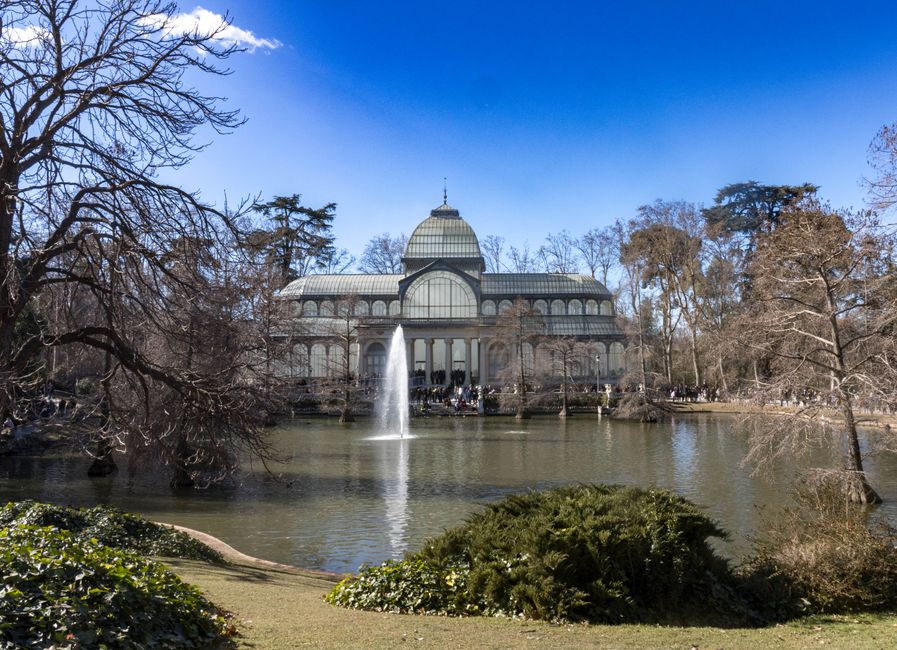 A large, glass-domed conservatory with a classical architectural style is situated across a pond in Crystal Palace Park on a bright, sunny day. The pond features a central fountain that sprays water high into the air, surrounded by bare deciduous trees and evergreen bushes. The conservatory's structure includes multiple arches, a prominent central dome, and a spire at the top. The area around the pond has pathways with groups of people visible, indicating visitors enjoying the park. The scene is well-lit with clear blue sky and natural light illuminating the landscape, with some shadows cast by the trees and structures. This setting is associated with house removals and moving services, illustrating a picturesque backdrop for furniture transport, packing, and loading processes involved in home relocation, as handled by Man With a Van Crystal Palace.