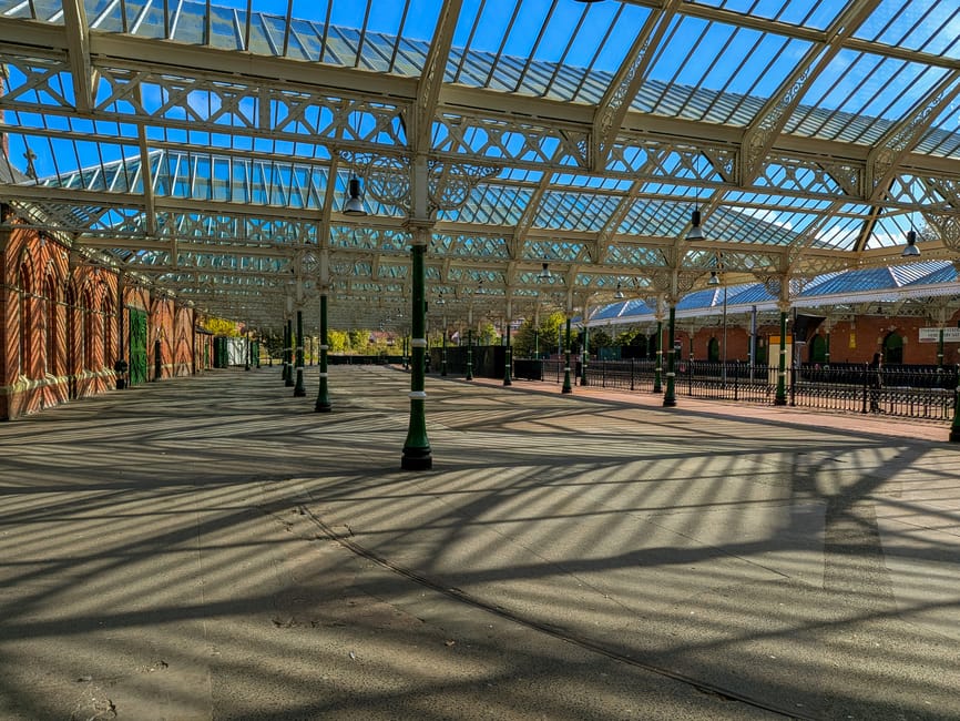 An empty outdoor area at Crystal Palace Station with a glass and metal canopy overhead, supported by ornate metal columns. The ground is paved with concrete, and long shadows from the canopy's iron framework stretch across the surface. To the left, a red brick wall with green doors is visible, while on the right, black metal fencing encloses the platform area. In the background, there is a small building with a blue roof and window, and trees are visible beyond the station. The lighting is bright, indicating a clear, sunny day. This setting exemplifies a typical home relocation or moving logistics environment, where furniture, boxes, or packing materials could be loaded or unloaded for transportation by services such as Man With a Van Crystal Palace, providing efficient and accessible moving or furniture transport solutions.