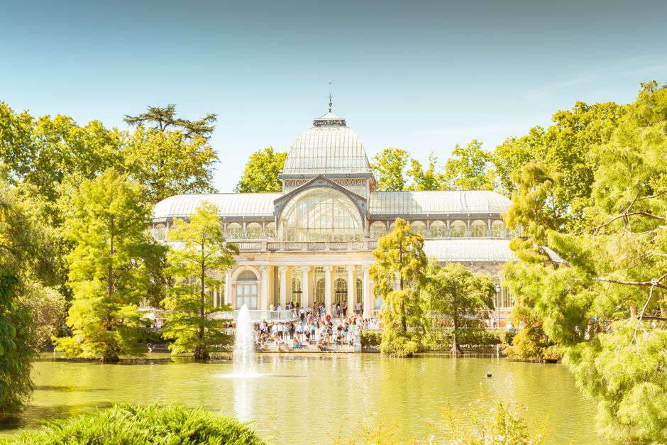 A large glass greenhouse with a domed roof and ornate architectural details stands behind a pond filled with calm water, reflecting the surrounding greenery. Tall trees with bright green foliage surround the pond and the greenhouse, creating a lush natural setting. In front of the greenhouse, a group of people can be seen walking and gathering on a paved area, suggesting visitors or clients during the moving or packing process. A small water fountain is positioned in the pond, adding motion to the scene. The bright daylight illuminates the scene, enhancing the vibrant colors of the trees and the building's glass and metal framework. This setting appears to be an outdoor location adjacent to a residential or historic property, with surroundings compatible with house removals or home relocation activities, which a professional removal service like Man With a Van Crystal Palace might facilitate.
