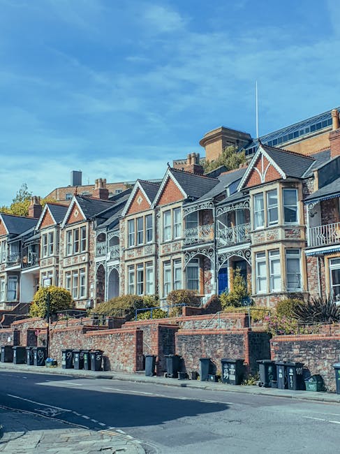 A row of Victorian-style terraced houses with ornate ironwork balconies and bay windows, situated along a quiet residential street with a brick retaining wall and small landscaped gardens in front. The houses are built with brick and feature gabled roofs, some with decorative trim. Above the houses, a clear blue sky with a few wispy clouds is visible, and in the background, there are taller buildings and a hillside. The street is lined with parked bins and a parked van, indicating ongoing home relocation or moving activity. The image captures a typical neighbourhood in the SE19 area, highlighting architecture and street scene relevant to house removals and furniture transport services provided by Man With a Van Crystal Palace.